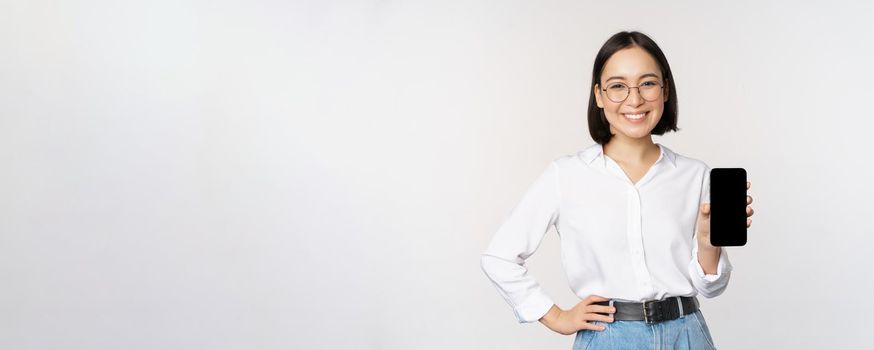 Smiling korean woman showing smartphone screen, demonstrating mobile application, standing over white background.