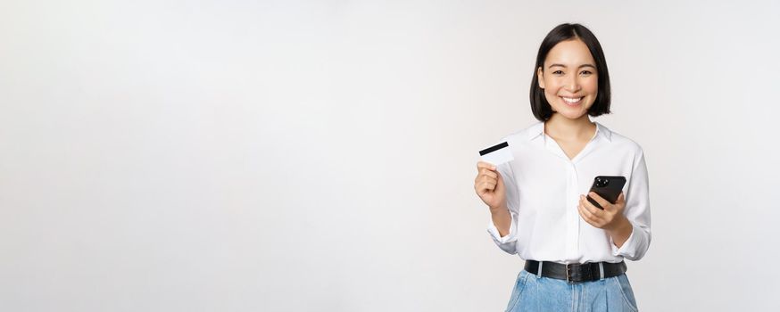 Online shopping concept. Image of young asian modern woman holding credit card and smartphone, buying with smartphone app, paying contactless, standing over white background.