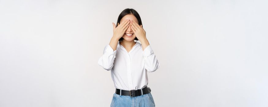 Image of young asian woman close her eyes and smiles, waits for surprise, anticipates, stands over white background.