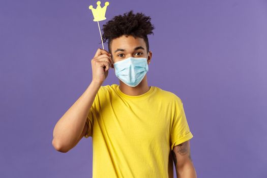 Social-distancing, coronavirus, health concept. Portrait of upbeat, happy hispanic man in medical face mask, holding small paper crown above head, staying home during quarantine, playing games.