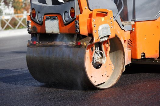Close-up view of steam roller working on new asphalt road at summer daylight. Water stream splashing on rolling barrel.