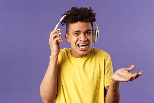 What do you want, I am in headphones. Portrait of confused annoyed young man shrugging, take-off earphone to hear what person asked while he was listening music, purple background.