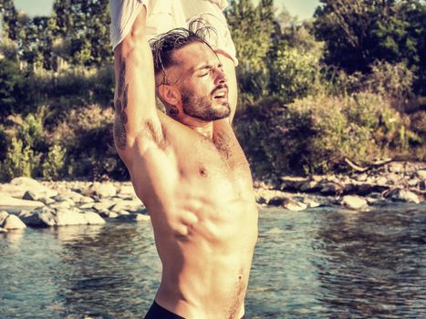 Handsome Muscular Shirtless Hunk Man Undressing, Removing T-Shirt Outdoor Next to River. Showing Healthy Muscle Body