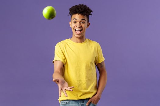 Holidays, vitamins and vacation concept. Portrait of handsome upbeat young male student asking friend something eat, catching apple and smiling happy, standing purple background.