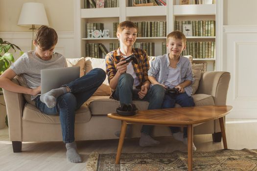 Three brother boys sit at home on the sofa in the sunlight and emotionally play computer and video games with joysticks in their hands