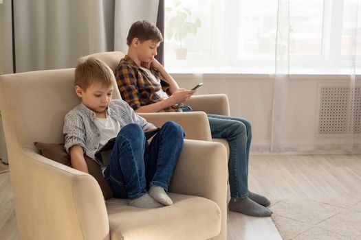 Two boys, a child and a teenager, sit in beige armchairs, in a room, by the window during the day, play their gadgets. Copy space