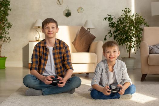 Portrait of two smiling boys sits on the floor in a room playing video games with joysticks. Close up