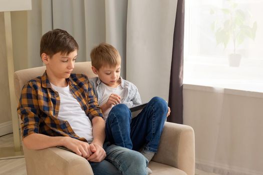 Two boys, a child and a teenager, sit in beige armchair, in a room, by the window during the day, using mobile device. Copy space