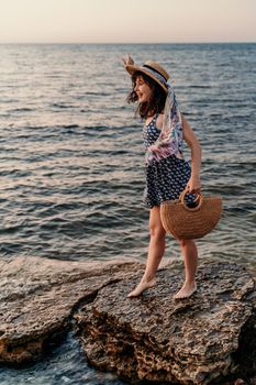 A woman in a dress, hat and with a straw bag is standing on the beach enjoying the sea. Happy summer holidays.