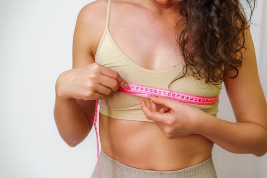 Cropped view of slim woman measuring breasts with tape measure at home, close up. Unrecognizable European woman checks the result of a weight loss diet or liposuction indoors. Healthy lifestyle..