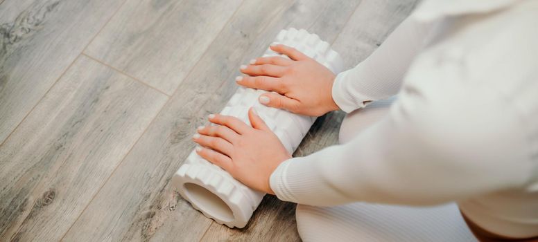 Adult athletic woman, in white bodysuit, performing fascia exercises on the floor - caucasian woman using a massage foam roller - a tool to relieve tension in the back and relieve muscle pain - the concept of physiotherapy and stretching training.