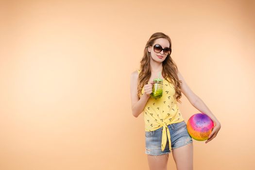 A woman in bright clothes, a yellow T-shirt and denim shorts, sunglasses holds a ball and cold lemonade in her hands, on an isolated background.