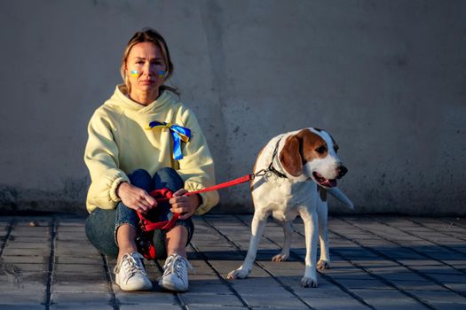 Ukrainian refugee woman with national flag on cheek sitting underground on floor with dog and crying