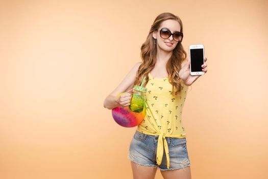 A woman in bright clothes, a yellow T-shirt and denim shorts, sunglasses holds a ball and cold lemonade in her hands, on an isolated background.