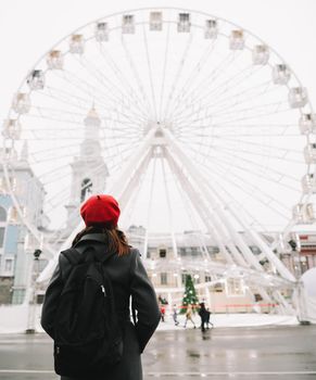 Young woman traveler in red hat and black coat looking at the ferris wheel in Kyiv, Ukraine. Outdoors photo of a girl on the background of modern city architecture.