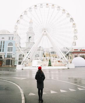Young woman traveler in red hat and black coat looking at the ferris wheel in Kyiv, Ukraine. Outdoors photo of a girl on the background of modern city architecture.