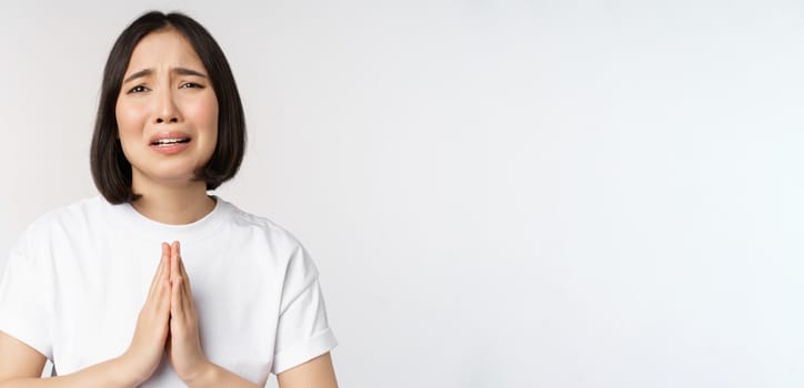 Desperate crying asian woman begging, asking for help, pleading and say please, standing in white t-shirt over white background.