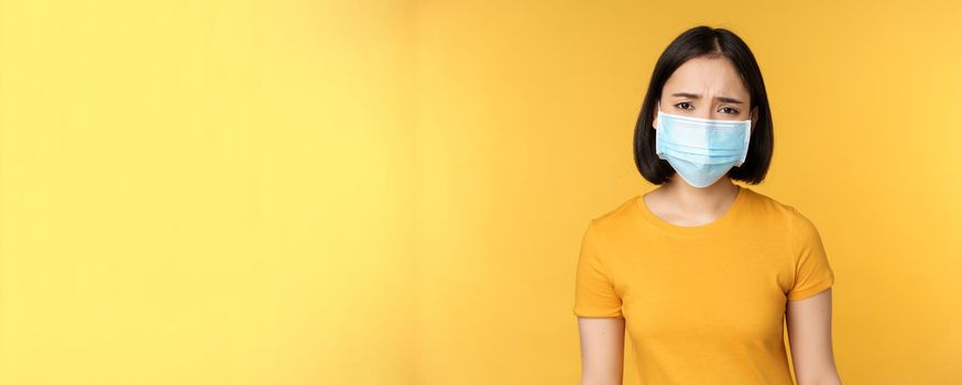 Image of sad and disappointed asian woman in medical mask, frowning and looking displeased, standing against yellow background.