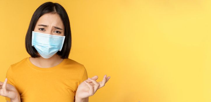 Close up portrait of confused asian woman in medical face mask, shrugging shoulders and looking puzzled, standing against yellow background.