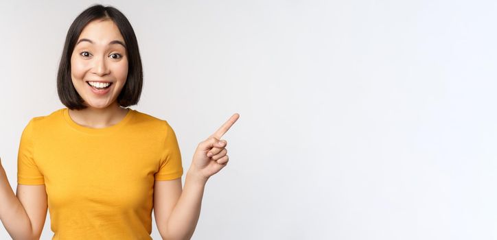 Cute asian girl pointing fingers sideways, showing left and right promo, two choices, variants of products, standing in yellow tshirt over white background.