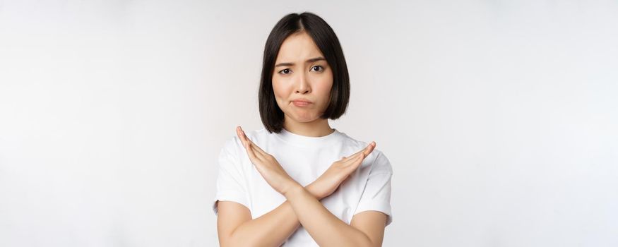 Portrait of asian korean woman showing stop, prohibition gesture, showing arm cross sign, standing in tshirt over white background. Copy space