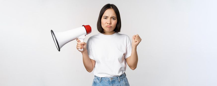 Angry asian girl activist, holding megaphone and looking furious, protesting, standing over white background. Copy space