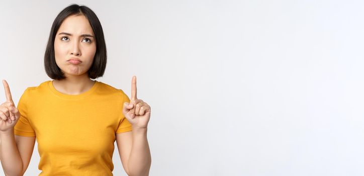 Disappointed asian woman looking, pointing fingers up with angry moody face expression, standing in yellow tshirt over white background.