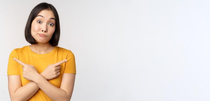 Complicated asian girl pointing fingers sideways, showing left and right choices, staring indecisive clueless, standing over white background. Copy space