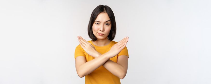 Portrait of asian woman looking serious and angry, showing stop prohibit gesture, taboo sign, forbidding smth, standing in yellow tshirt over white background.