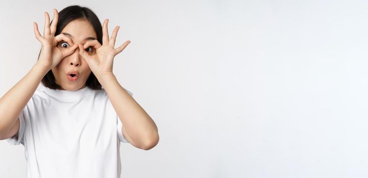 Funny young asian woman, korean girl making eyes glasses gesture, looking happy at camera, standing over white background.