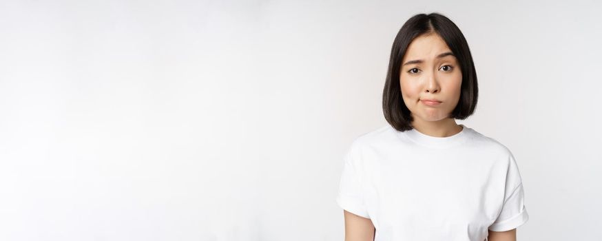 Image of young asian brunette woman in white t-shirt, smirk and look with doubt, hesitating, standing against white background. Copy space