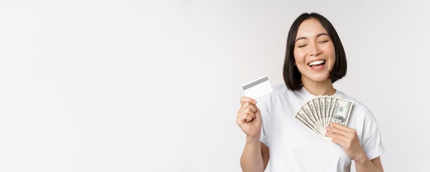 Portrait of asian woman smiling, holding credit card and money cash, dollars, standing in tshirt over white background.