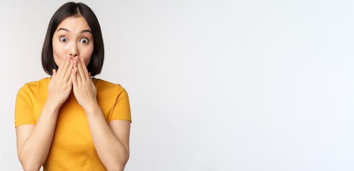 Surprised asian woman gasping, cover mouth with hands and looking amazed at camera, wearing yellow t-shirt, standing over white background.