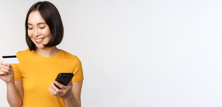 Beautiful smiling asian girl using credit card and mobile phone, paying online on smartphone, standing in yellow tshirt over white background.