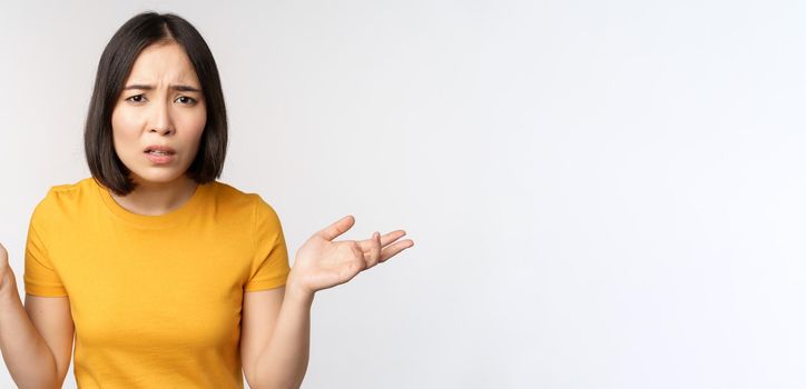 Frustrated, confused asian woman cant understand smth, looking clueless, shrugging shoulders, standing in yellow tshirt over white background.