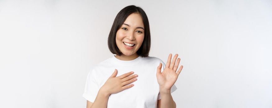 Portrait of beautiful korean girl raising hand, introduce herself, put hand on heart, greeting, standing over white background.