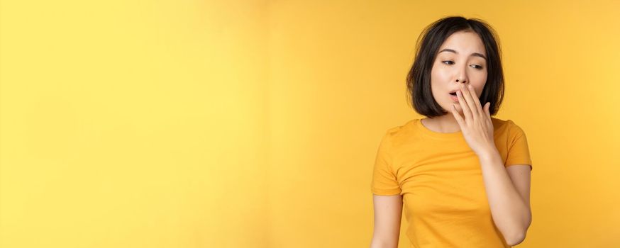 Image of young asian girl student, yawning, feeling tired, standing sleepy agaisnt yellow background. Copy space