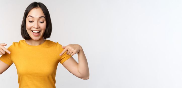 Happy asian girl pointing fingers down, looking at announcement, banner or advertisment, standing in yellow tshirt over white background.