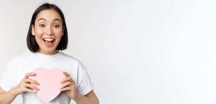 Valentines day. Happy asian girl receive romantic gift, holding heart shaped box and smiling excited, standing over white background.