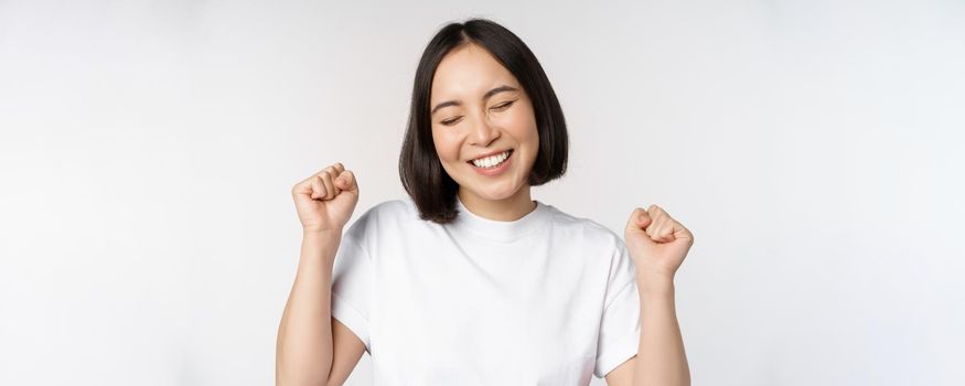 Dancing asian girl celebrating, feeling happy and upbeat, smiling broadly, standing over studio white background.