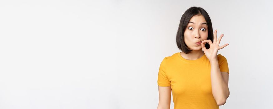 Cute asian girl seal lips, zipping mouth with finger, promise to keep secret, taboo gesture, standing in yellow tshirt over white background.