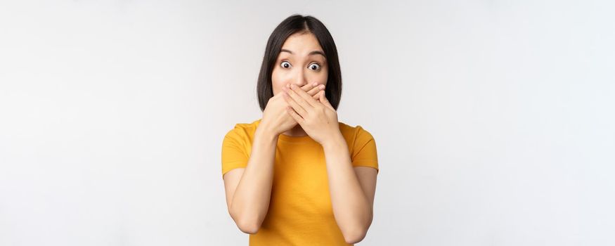 Shocked asian woman cover mouth with hands, looking startled with speechless face expression, standing in yellow tshirt against white background.