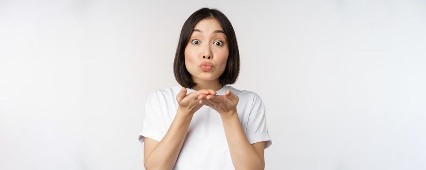 Lovely young asian woman, sending air kiss and looking coquettish at camera, standing in tshirt over white background.