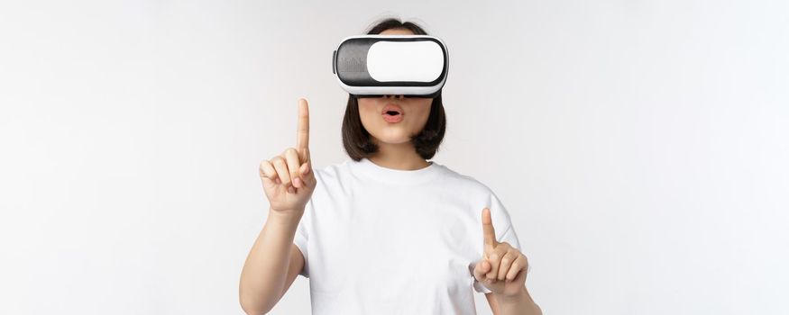 Potrait of asian woman in virtual reality glasses, pointing, choosing smth in VR headset, standing over white background.