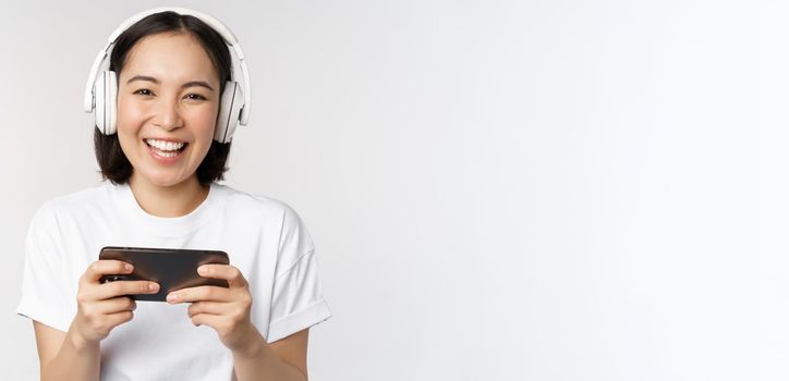 Happy asian girl gamer, playing on mobile phone, watching on smartphone, wearing headphones, standing over white background.