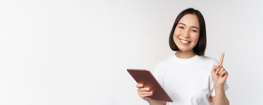 Enthusiastic asian woman with tablet, raising finger and looking amazed, pointing up, standing over white background.