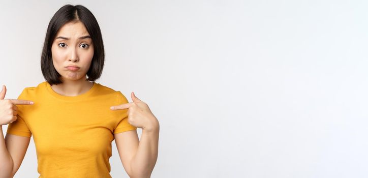 Confused asian woman pointing at herself, looking in disbelief, standing over white background. Copy space