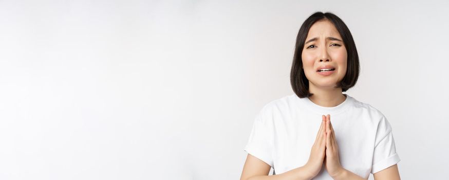 Desperate crying asian woman begging, asking for help, pleading and say please, standing in white t-shirt over white background.