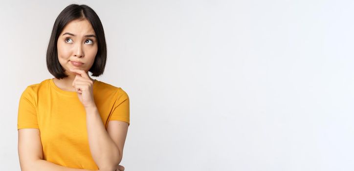 Image of thinking asian woman, looking aside and pondering, making decision, standing in yellow tshirt over white background.