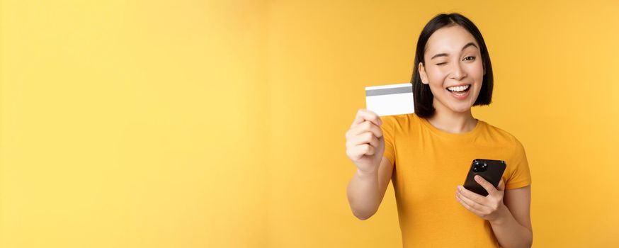 Joyful asian girl smiling, showing credit card and smartphone, recommending mobile phone banking, standing against yellow background.